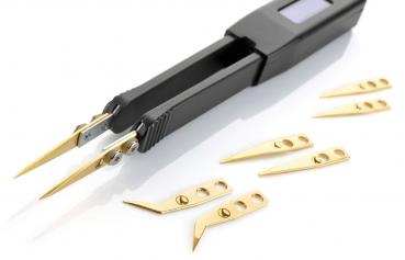 Auf dem Bild sind eine schwarze elektronische Pinzette mit zwei spitzen Greifarmen und mehrere kleine, goldene Schneideblätter zu sehen. Die Blätter sind unterschiedlich geformt.