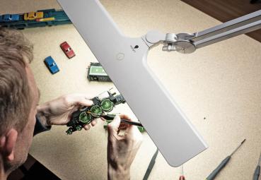 A person is working intently on a small model. In front of them is a bright lamp. Around them, small toy cars and tools are scattered.