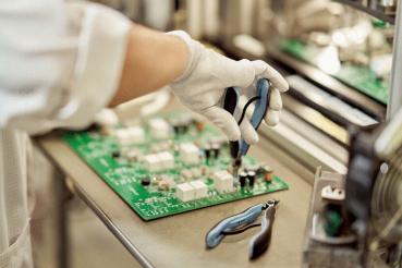 Eine Hand, die einen kleinen, blauen Zangenwerkzeughält, arbeitet an einer grünen Leiterplatte mit verschiedenen Bauteilen, wie Widerständen und Chips, auf einem Tisch.