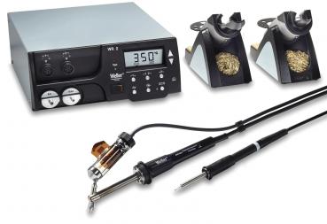 The image shows a soldering station with a digital display. There are two soldering irons in holders, along with cables and controls for adjusting the temperature.