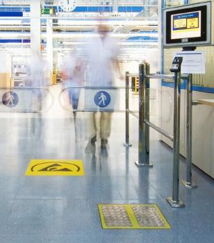 In the image, you can see a modernly designed entrance area with turnstiles, yellow floor markings, and people who are hurriedly passing by in the background.