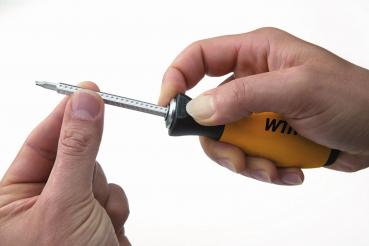 In the picture, there is a hand holding a screwdriver with a yellow handle. The tip of the screwdriver is pointing upwards, while a second hand carefully holds it steady.