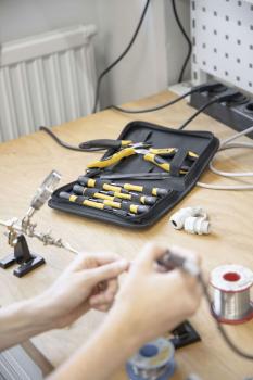 On a table, there are tools in a black case with yellow handles. One hand is holding a soldering iron, while the other works on a wire. In the background, cables and a holder are visible.