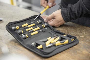 The picture shows a tool bag with several screwdrivers. The tools have gray handles with yellow accents. A hand is holding a screwdriver while the bag is on a table.