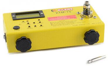 The image shows a yellow device that is rectangular. It has a digital display, several buttons, and two large, black knobs. Next to it lies a small silver object.
