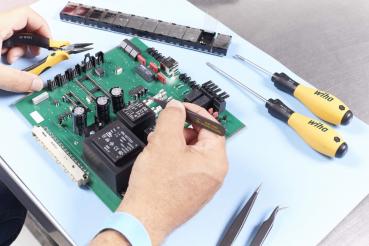 In the image, you can see a manual assembly or repair of a green circuit board with many electronic components. Next to it, there are screwdrivers and pliers.
