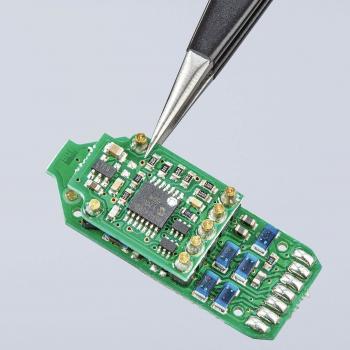 The image shows a green circuit board in a technical environment. A pair of tweezers is holding a small component in place. The surface is equipped with various components and connections.