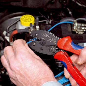A hand holds a wire cutter with red and blue handles while working on a blue cable leading to a car component. In the background, a yellow container is visible.