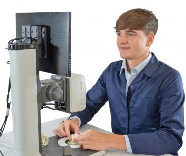 Ein junger Mann sitzt an einem Tisch, vor ihm ein Monitor und ein Gerät zur Vergrößerung. Er schaut konzentriert auf einen kleinen Gegenstand, den er mit einer Hand hält.