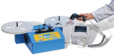 Das Bild zeigt ein blaues Gerät mit zwei runden Scheiben oben und einem Drucker daneben. Eine Hand hält einen Barcode-Scanner. Das Gerät hat ein Display und Tasten zur Bedienung.