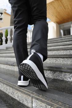 Eine Person geht die Treppe hinauf, trägt schwarze Sportchuhe mit weißen Details und schwarze Hose. Im Hintergrund sind gelbe und graue Wandflächen sichtbar.