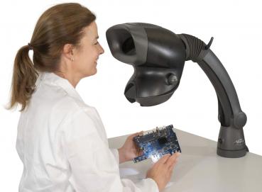 Eine lächelnde Frau in einem weißen Laborkittel hält eine elektronische Platine. Vor ihr steht ein Vergrößerungsgerät mit einem beweglichen Arm, das auf die Platine gerichtet ist.