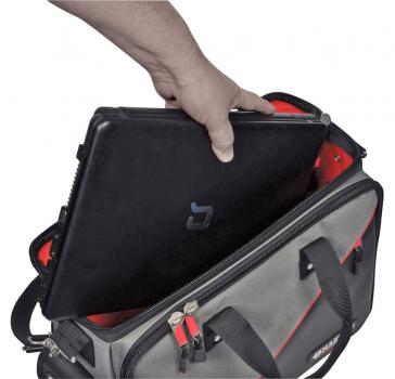 A hand places a black laptop into a gray bag with red lining. The bag has two handles and an open zipper.