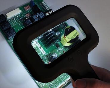 Das Bild zeigt eine Hand, die eine Lupe über eine grüne Leiterplatte hält. Die Lupe beleuchtet einen Bereich mit verschiedenen elektrischen Bauteilen, darunter ein schwarzes Element und ein gelbes.