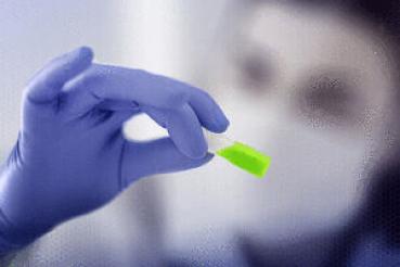 Ein Hand mit blauen Handschuhen hält ein kleines, leuchtend grünes Objekt. Im Hintergrund ist unscharf eine Person mit Mundschutz zu erkennen.