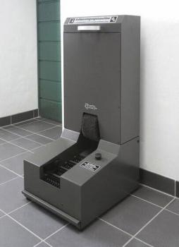 Das Bild zeigt eine graue Maschine zur Mülltrennung. Sie hat einen oberen Bereich mit einer Klappe und darunter eine breite Öffnung zur Abfallannahme, auf einem Fliesenboden.