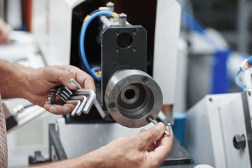 In the picture, hands are visible holding tools (Allen wrenches). In the background, a machine component can be seen, which is round and metallic. It is an industrial environment.