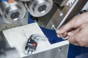 A craftsman uses an Allen wrench to work on a component of a machine. In the foreground, several Allen wrenches are lying on a workbench.