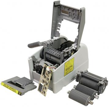 The image shows an open mechanical device with a gray casing. Inside, gears and a circuit board are visible. On the side, there is a small black block, presumably a part of the device.