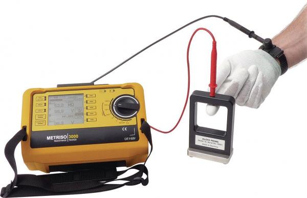 The image shows an electric measuring device in yellow with a screen and buttons. A cable connects it to a handheld device that has a metallic surface. A hand in a white glove is holding the handheld device.