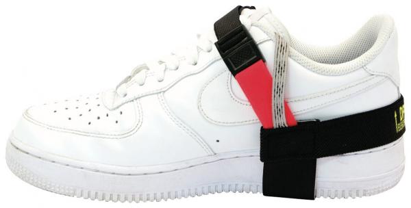 Das Bild zeigt einen weißen Sneaker der Marke Nike. Am Schuh ist ein schwarzes Band mit einer roten und einer gelben Markierung befestigt, das teilweise um den Schuh gewickelt ist.