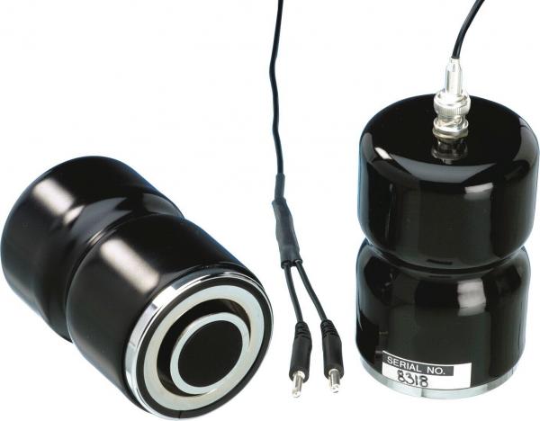 The image shows two black, round speakers with a glossy surface. They are of different heights and are connected by cables. One has a label with a serial number on it.