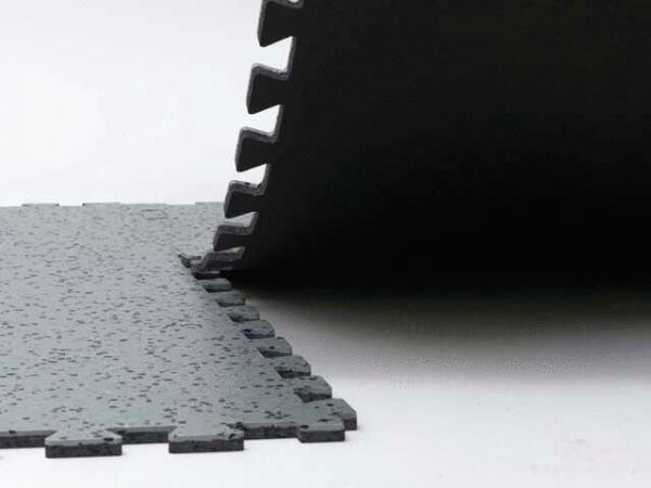 The image shows a black, textured surface made of thick mats stacked at one edge. One mat is slightly lifted, while the other is flat. The surface has a rough texture.