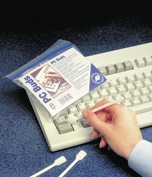 Auf dem Bild sieht man eine Hand, die einen leicht transparenten Beutel mit "PC Buds" hält. Daneben liegt eine weiße Tastatur und zwei Wattestäbchen auf einem blauen Untergrund.