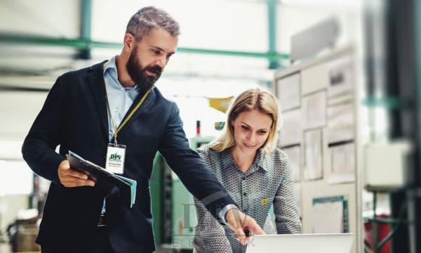 Ein Mann und eine Frau stehen in einem modernen Büro. Der Mann hält ein Tablet und zeigt auf einen Laptop, während die Frau aufmerksam darauf schaut. Beide wirken konzentriert und freundlich.