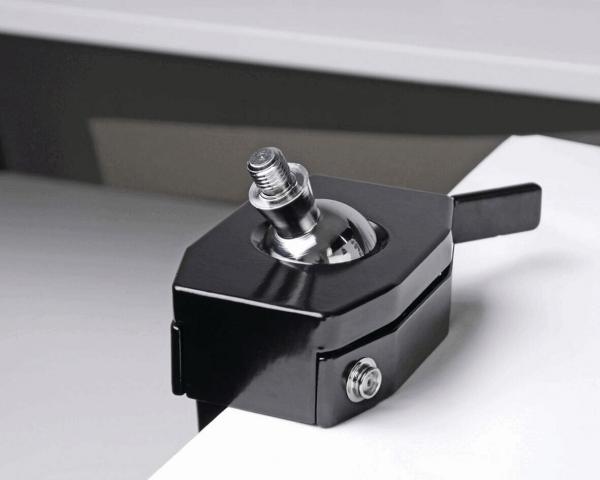 The image shows a black clamping mechanism with a silver, round head. The mechanism is positioned on the edge of a table and has a lever for tightening.