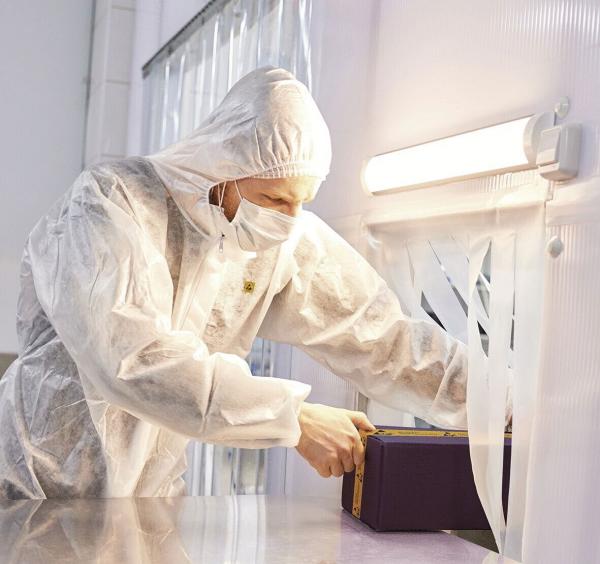 Eine Person in einem weißen Schutzhanzug und Maske arbeitet an einem Tisch. Sie öffnet eine lila Kiste. Der Raum wirkt steril, mit hellem Licht und durchsichtigen Vorhängen.