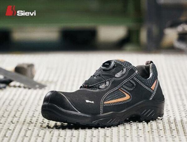 Das Bild zeigt einen schwarzen Arbeitsschuh mit runder Zehenkappe. Er hat orangefarbene Akzente und eine rutschfeste Sohle. Der Schuh ist auf einem grauen, strukturieren Untergrund platziert.