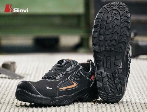Das Bild zeigt ein Paar schwarze Sicherheitsschuhe mit rutschfester Sohle. Sie sind leicht und haben orangefarbene Designelemente. Die Sohle hat ein tiefes Profil für besseren Halt.