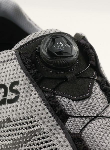 Das Bild zeigt einen grauen Sportschuh. Oben befindet sich ein schwarzes Schnürsystem mit einem runden Klickmechanismus. Der Schuh hat ein atmungsaktives Mesh-Material.