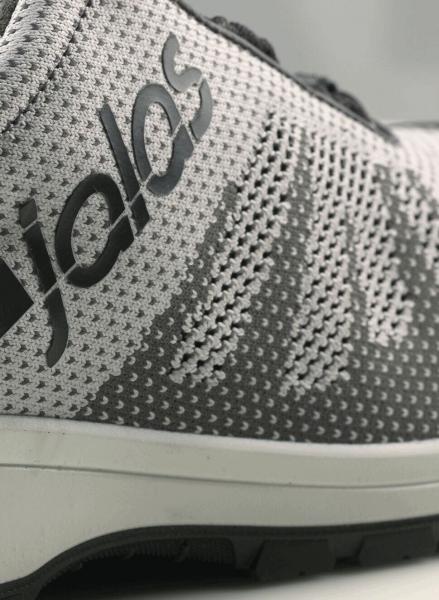 Das Bild zeigt einen sportlichen Schuh in Grau und Weiß mit einem modernen, strukturierten Obermaterial. Der Markenname ist seitlich in Schwarz aufgedruckt. Die Sohle ist dunkel und rutschfest.