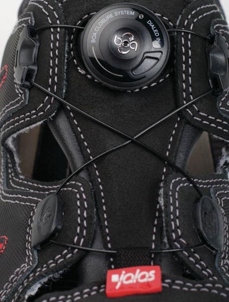 Das Bild zeigt einen schwarzen Schuh mit einem speziellen Schnürsystem. In der Mitte befindet sich eine runde Drehschließe, die die Schnürsenkel strafft. Auf der Sohle steht "jalas".