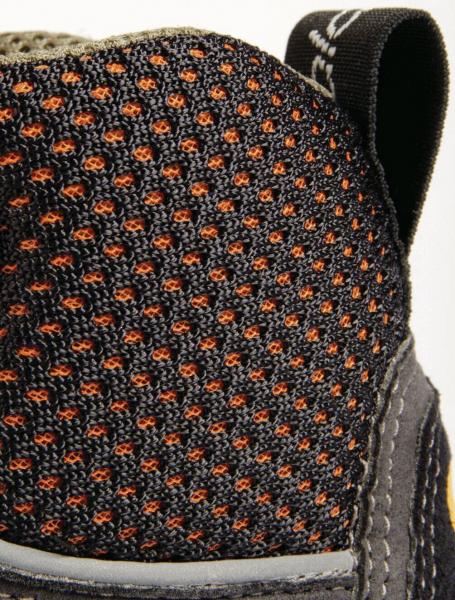 The image shows the upper part of a shoe. The surface is made of black mesh fabric with fine, orange dots. A thin, gray border runs along the bottom. A loop is visible at the top.
