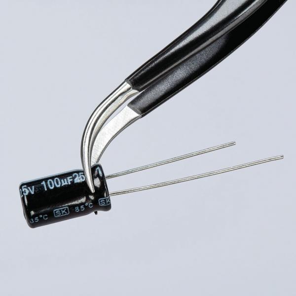 The image shows a pair of tweezers holding a cylindrical, black capacitor. The capacitor has two metal terminals and markings in white and gold.