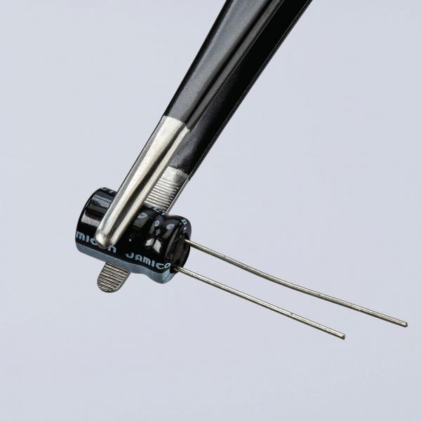 The image shows a pair of pliers holding a small, round, black component with two protruding wires. The component has a smooth surface and bears an inscription.