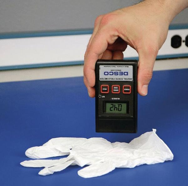 Eine Hand hält ein Messgerät über weißen Einweghandschuhen. Das digitale Display zeigt eine Zahl an, während der Hintergrund blau ist.