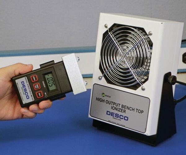 Das Bild zeigt einen ionisierenden Luftreiniger mit einem Ventilator. Jemand hält ein Messgerät in der Hand, das möglicherweise die Ionisierung misst. Beide Geräte stehen auf einem Tisch.