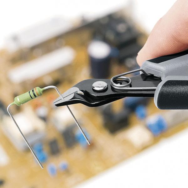 Auf dem Bild sieht man eine Hand, die eine Zange hält, die einen Widerstand greift. Im Hintergrund ist eine Leiterplatte mit verschiedenen elektronischen Bauteilen unscharf zu erkennen.