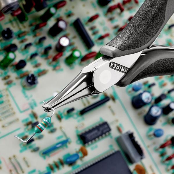 The image shows a pair of pliers holding a small electronic component, while in the background a complex circuit board with colorful components is visible.