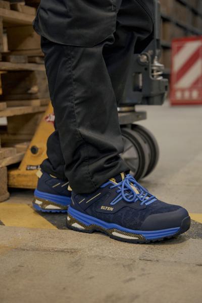 Das Bild zeigt einen stehenden Mann in einer dunklen Hose, der Sicherheitsschuhe in Blau und Schwarz trägt. Der Hintergrund ist ein Lager mit Holzpaletten.