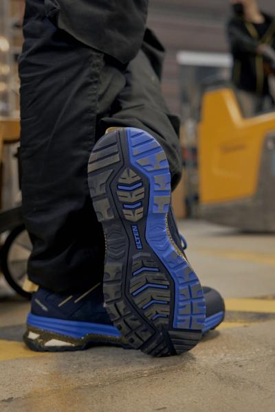 Das Bild zeigt die Unterseite eines blauen Schuhs auf einem Boden. Die Sohle hat ein markantes, rutschfestes Profil mit verschiedenen Mustern und Linien, die für guten Halt sorgen.