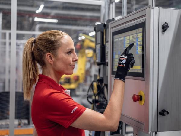 A woman in a red shirt is operating a touchscreen on a machine. She is wearing a black glove and looking intently at the screen. Robots are visible in the background.