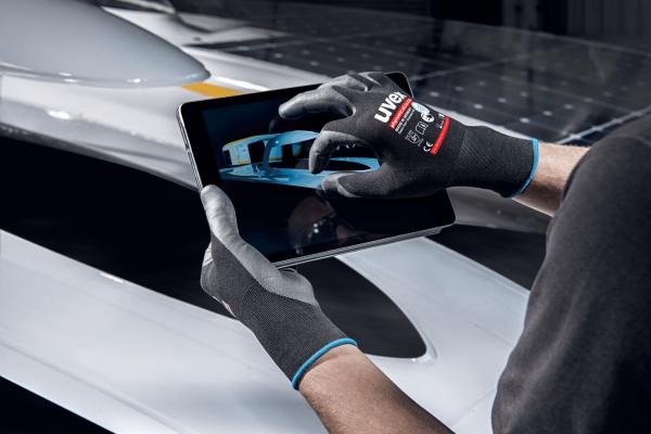 A person is wearing gray gloves and holding a tablet displaying an image of a vehicle. The background is dark and shows parts of a car.
