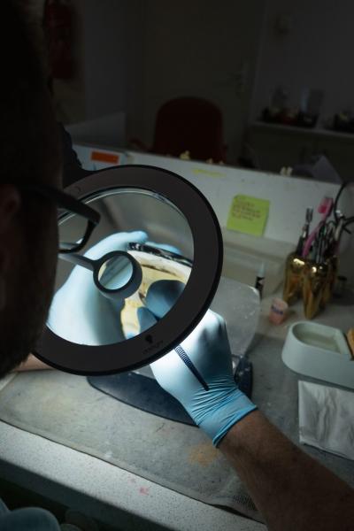 Eine Person mit blauen Handschuhen hält ein Objekt unter einer Lupe. Der Arbeitsplatz hat verschiedene Werkzeuge und Utensilien. Im Hintergrund sind unscharfe Möbel und einige farbige Notizzettel.