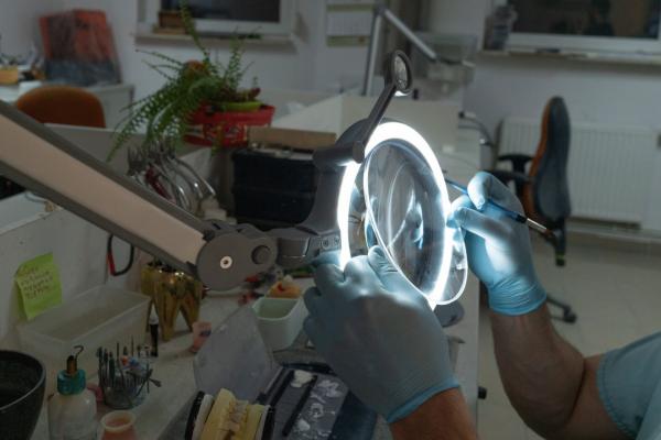 Eine Person mit blauen Handschuhen arbeitet mit einem Werkzeug unter einer vergrößernden Lampe. Der Arbeitsplatz ist ordentlich und enthält verschiedene Werkzeuge und Materialien.