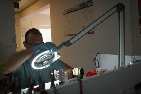 Der Bildausschnitt zeigt eine Person mit Handschuhen, die konzentriert unter einer hellen, runden Arbeitslampe an einem Tisch arbeitet. Im Hintergrund ist eine Wand mit einem Kalender sichtbar.